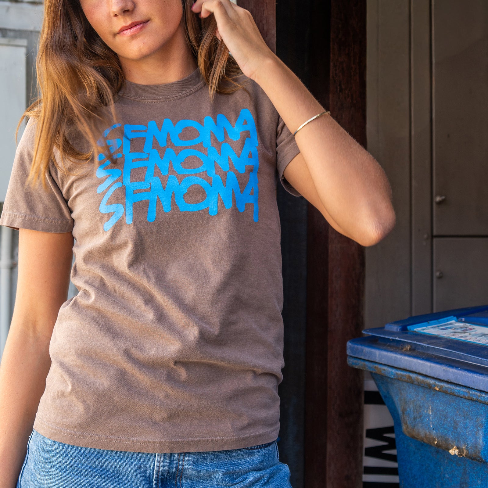 A dark gray short-sleeve crewneck t-shirt featuring the word 'SFMOMA' printed in a bright blue stylized stacked graphic on the chest, displayed flat against a white background.
