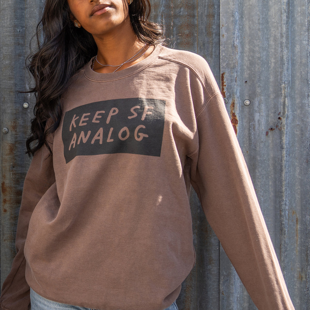 A brown ribbed crewneck sweatshirt featuring a black rectangular graphic on the chest with the text 'KEEP SF ANALOG' in a handwritten font, displayed flat against a white background.