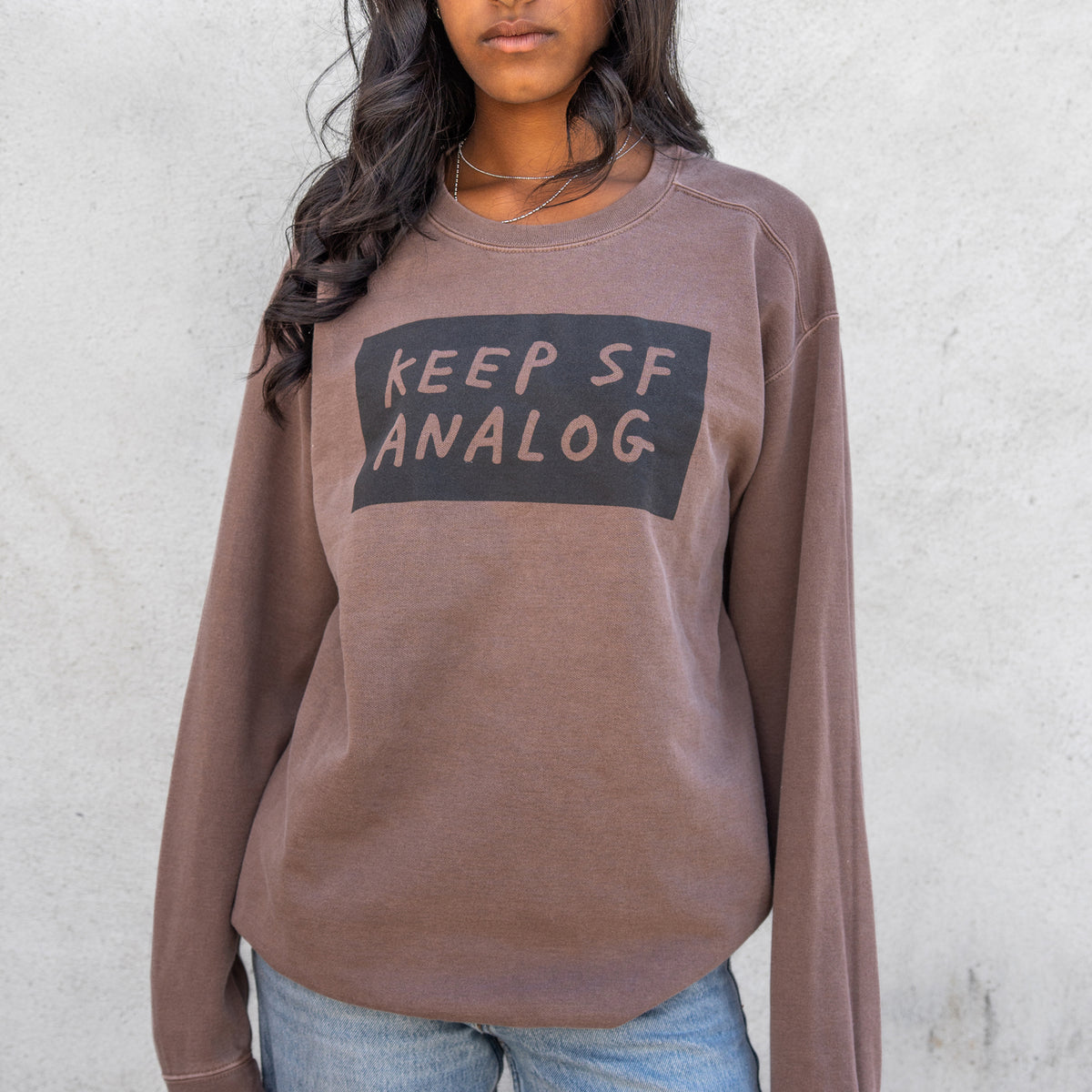 A woman wearing a brown long-sleeve sweatshirt with a black graphic box on the chest that reads 'KEEP SF ANALOG' in beige letters, paired with blue jeans against a light gray concrete wall.