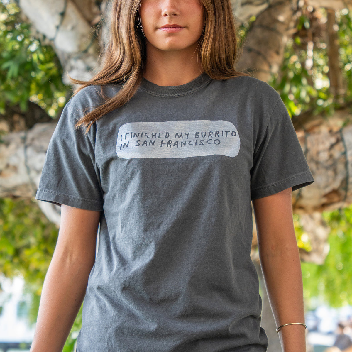 A woman wearing a dark gray t-shirt with a white graphic box containing the text 'I FINISHED MY BURRITO IN SAN FRANCISCO' in gray lettering, set against a blurred outdoor background of trees.
