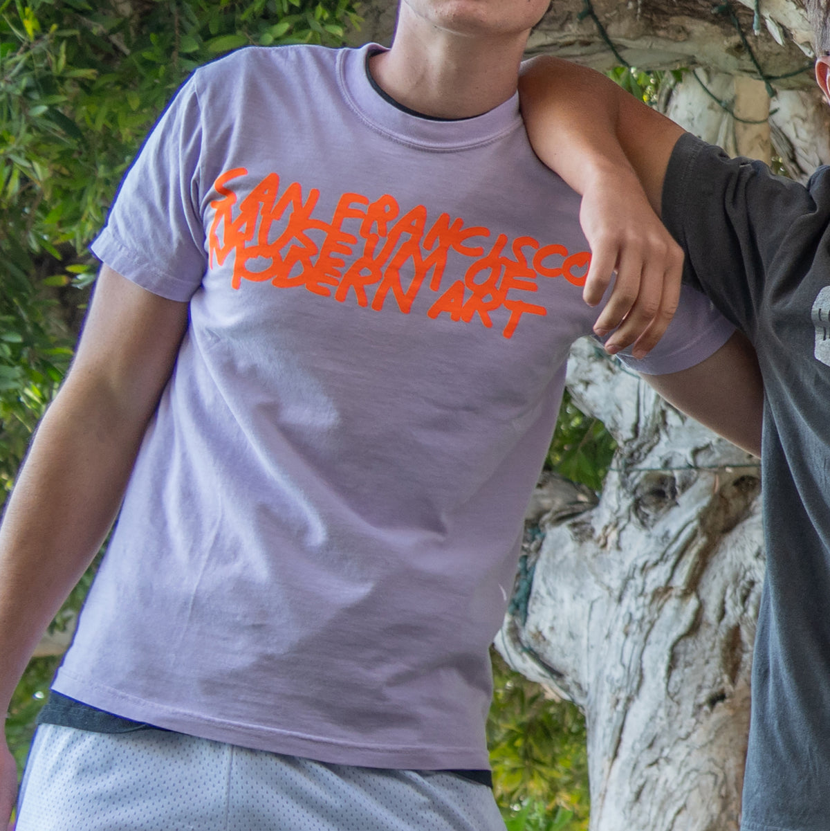 A person wearing a light purple t-shirt with orange text that reads 'SAN FRANCISCO MODERN ART' printed across the chest, standing outdoors next to a tree trunk.
