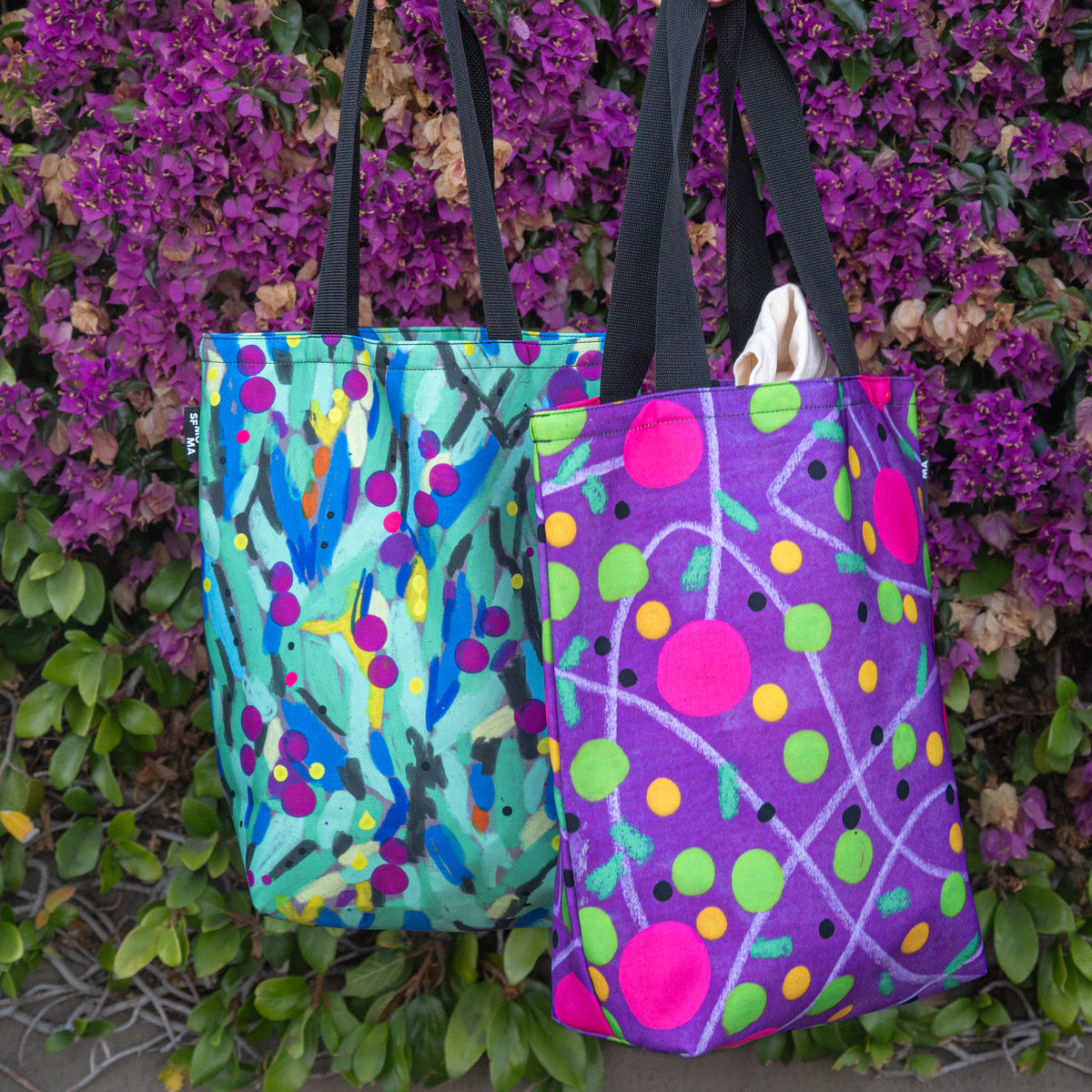 Two colorful canvas tote bags hanging in front of a wall of purple flowers and green leaves. The bag on the left is turquoise with abstract brushstrokes and colorful dots.