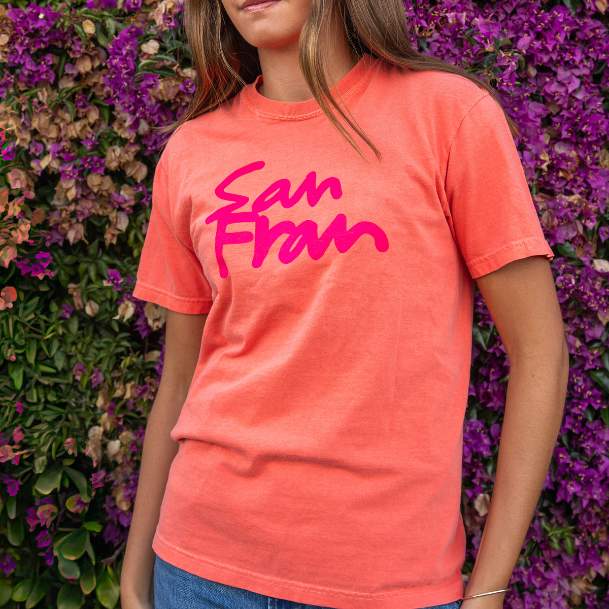 A bright coral orange crewneck t-shirt featuring the words 'San Fran' printed across the chest in a vibrant pink script font, displayed flat against a solid white background.