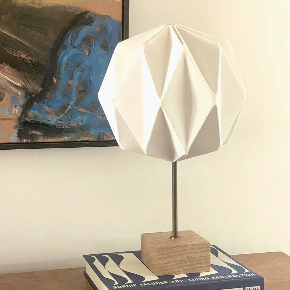 A modern table lamp with a white faceted geometric shade mounted on a square wooden base and thin metal stem, placed on top of a book against a cream wall with a framed painting behind it.