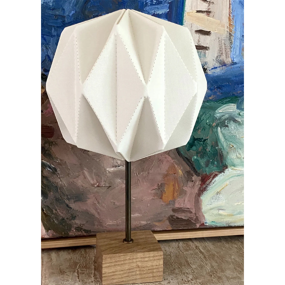 A modern table lamp with a white faceted geometric shade mounted on a square wooden base and thin metal stem, placed on top of a book against a cream wall with a framed painting behind it.