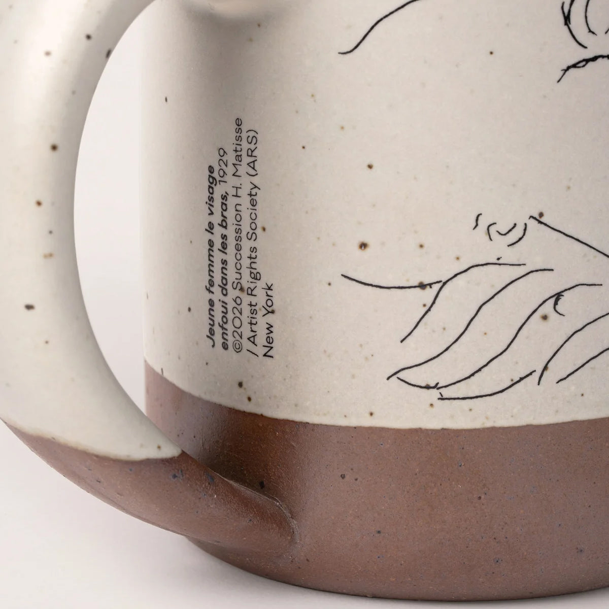 Close-up of a handmade ceramic mug showing a speckled cream upper body with black line art and text, a brown glazed base, and part of the handle on a white background.