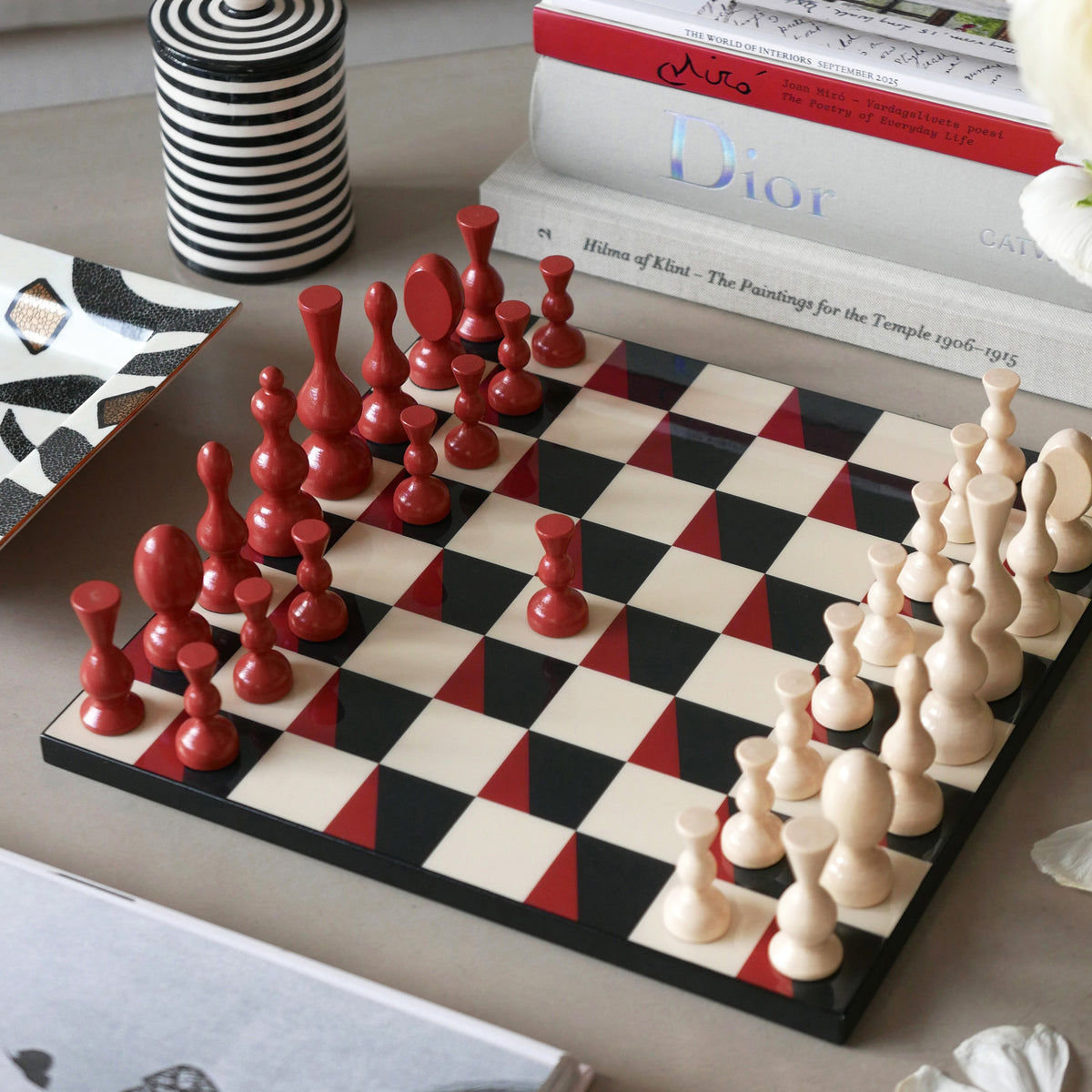 A wooden chess set with red and cream-colored pawns and pieces arranged on a checkered board, styled on a tabletop with books and a striped container in the background.