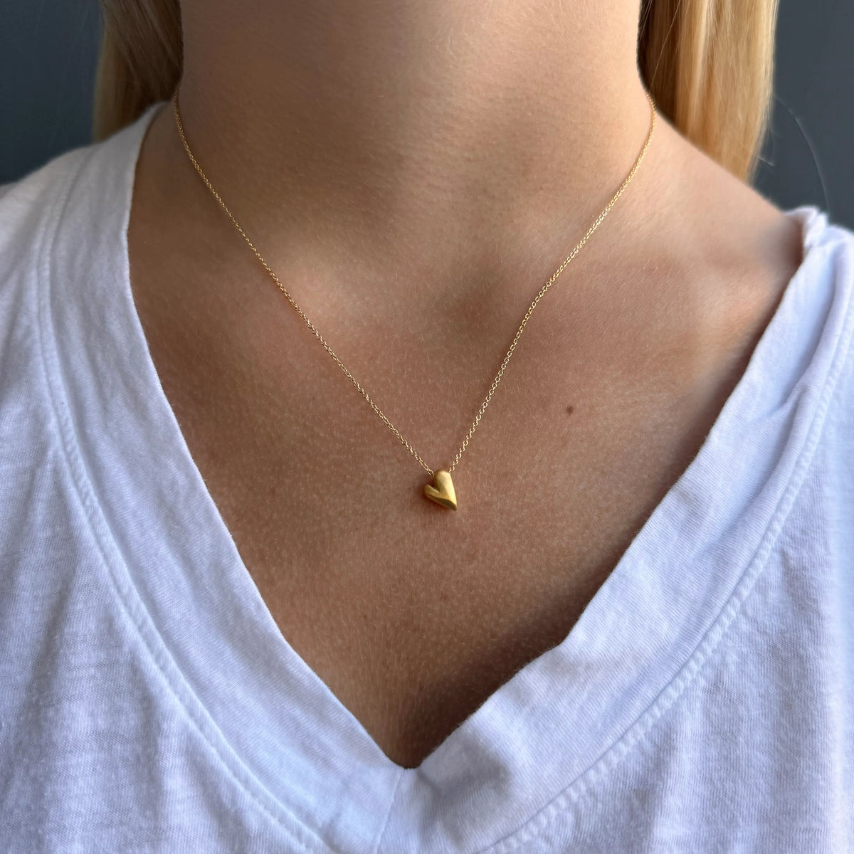 A close-up of a person wearing a delicate gold chain necklace with a small polished gold heart pendant, styled with a white v-neck t-shirt.