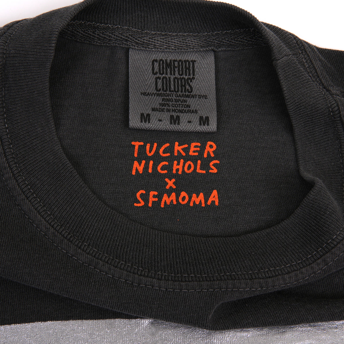 Close-up of the inside collar of a black Comfort Colors T-shirt with orange embroidered text reading 'TUCKER NICHOLS x SFMOMA'.