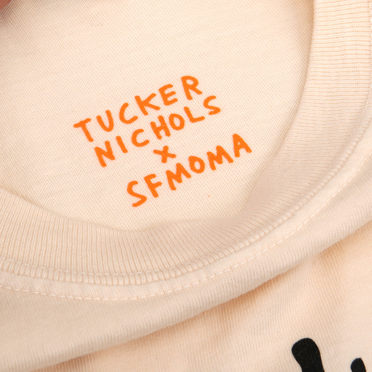 Close-up of a light biege t-shirt with orange printed text reading 'TUCKER NICHOLS x SFMOMA' on the inside collar.