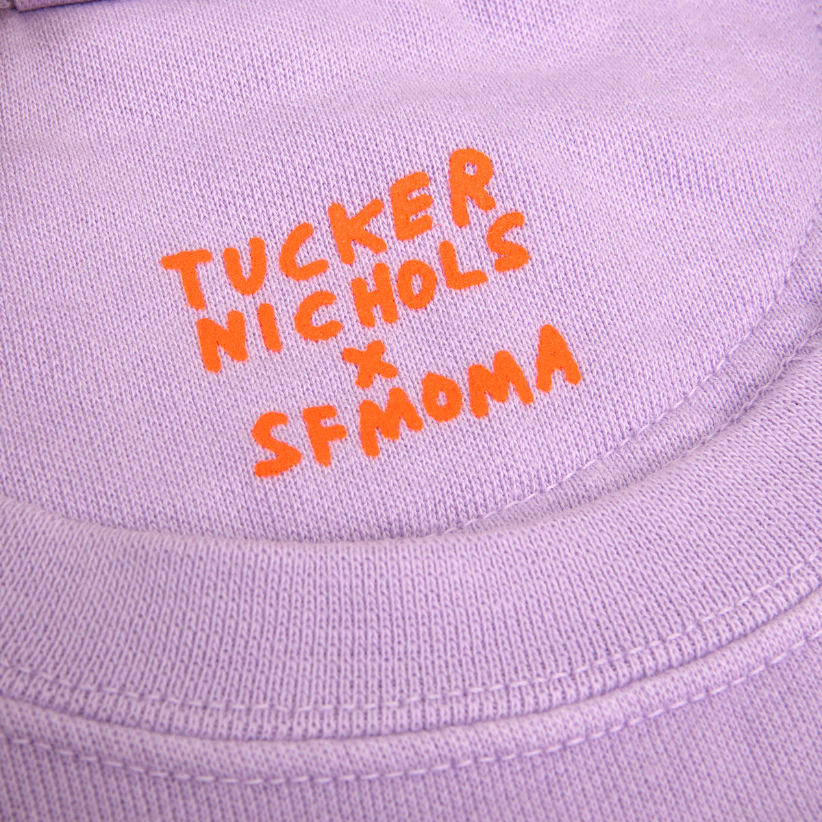 Close-up of a light lavender knit garment with bright orange  text reading 'TUCKER NICHOLS x SFMOMA' on the chest.