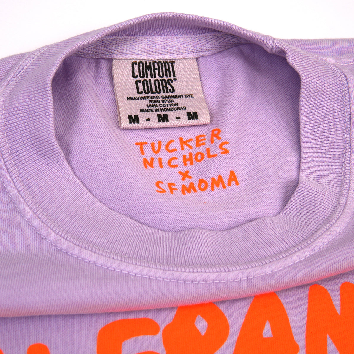Close-up of a lavender Comfort Colors t-shirt neck label with orange handwritten text reading 'TUCKER NICHOLS X SFMOMA' and orange graphic lettering at the bottom.