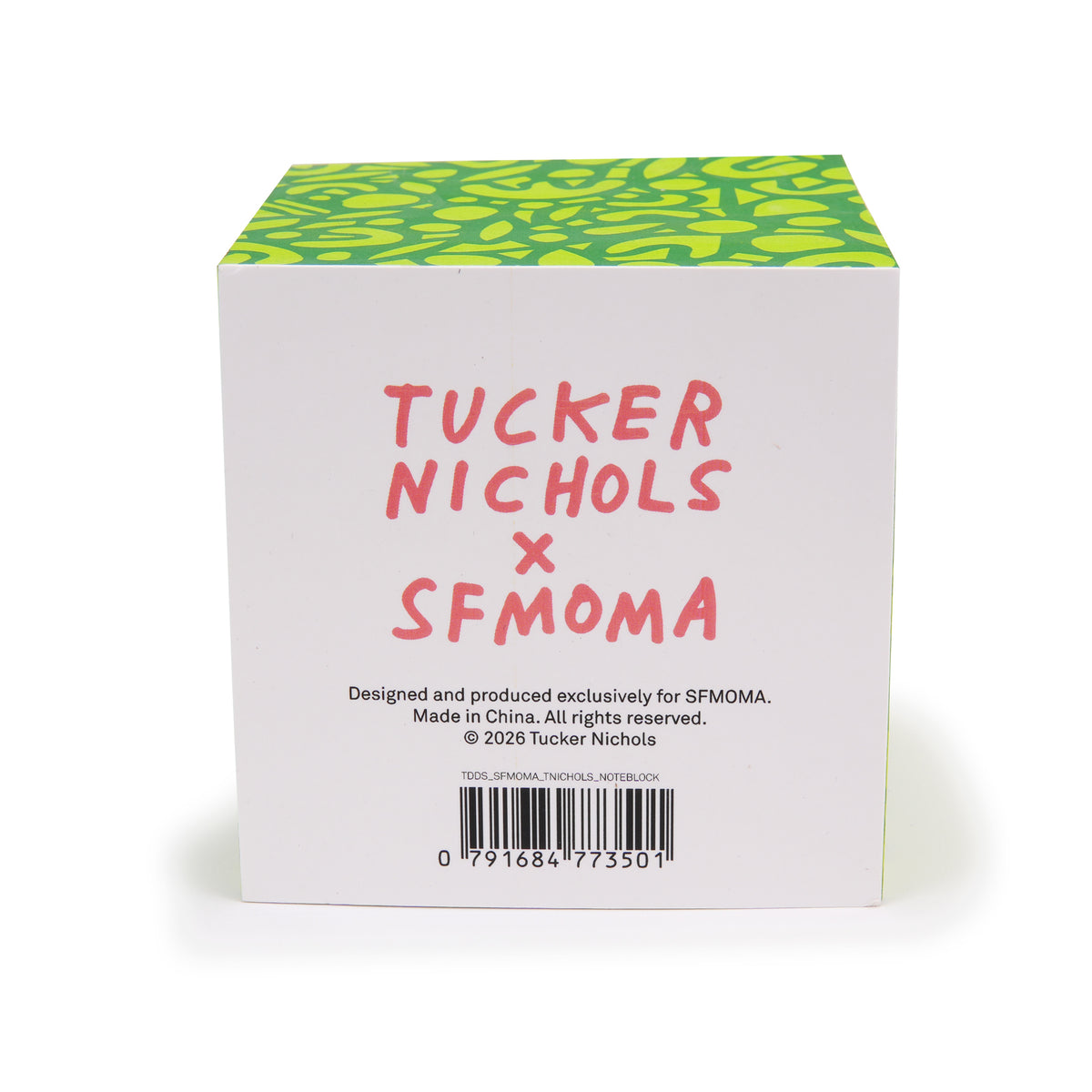 A square product box shown from the back, featuring a white face with pink handwritten text reading 'TUCKER NICHOLS x SFMOMA' and a green patterned top surface, photographed on a clean white background.