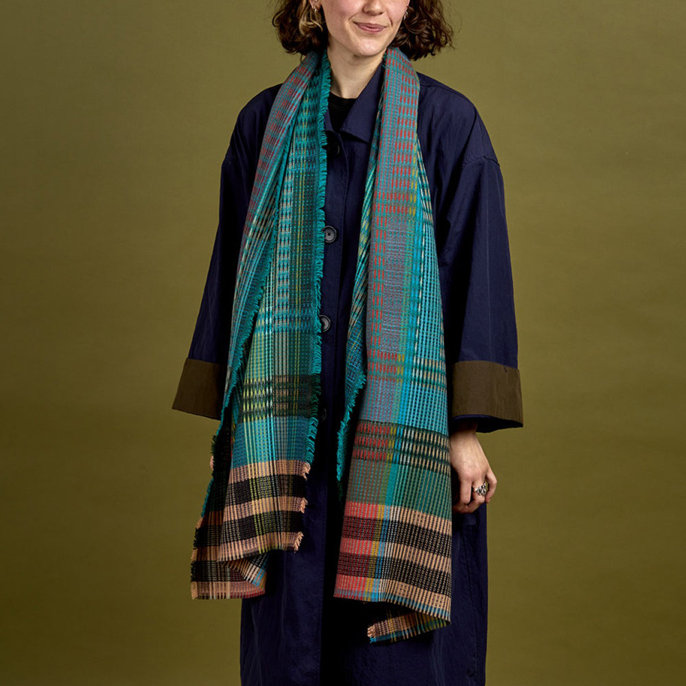 A model wearing a navy blue coat with brown turned-up cuffs and a long multicolor plaid scarf in shades of teal, green, orange, and brown, set against a solid olive green background.