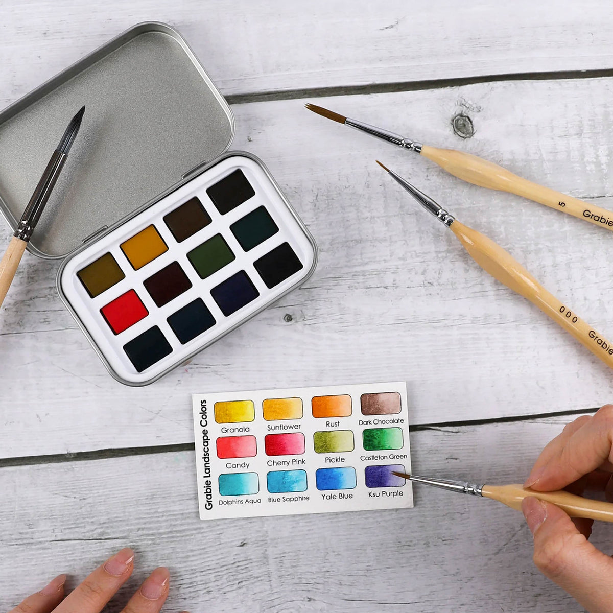 A watercolor paint palette in a silver metal tin, several paintbrushes, and a color chart labeled 'Gadla landscape colors' arranged on a white wood surface. Two hands are visible holding brushes near the palette.