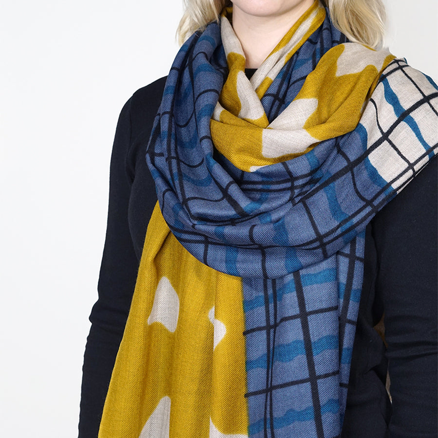 A close-up of a person wearing a patterned scarf with yellow, blue, and white sections, including plaid and abstract shapes, paired with a dark navy top against a plain white background.