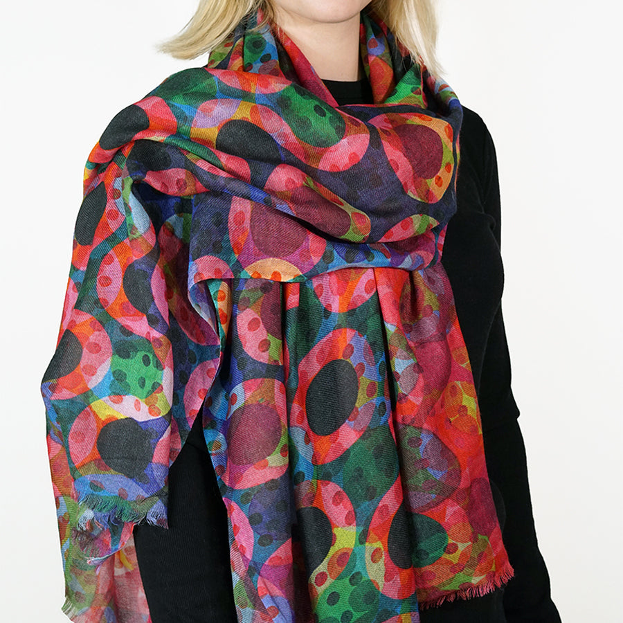 A multicolor abstract patterned scarf with pink, green, red, blue, and black circular motifs, worn by a model in a black top against a plain white background.