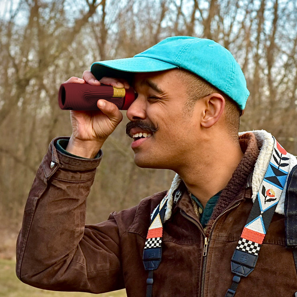 A man outdoors wearing a turquoise baseball cap, brown jacket, and patterned backpack strap, looking through a small red binocular-like device.