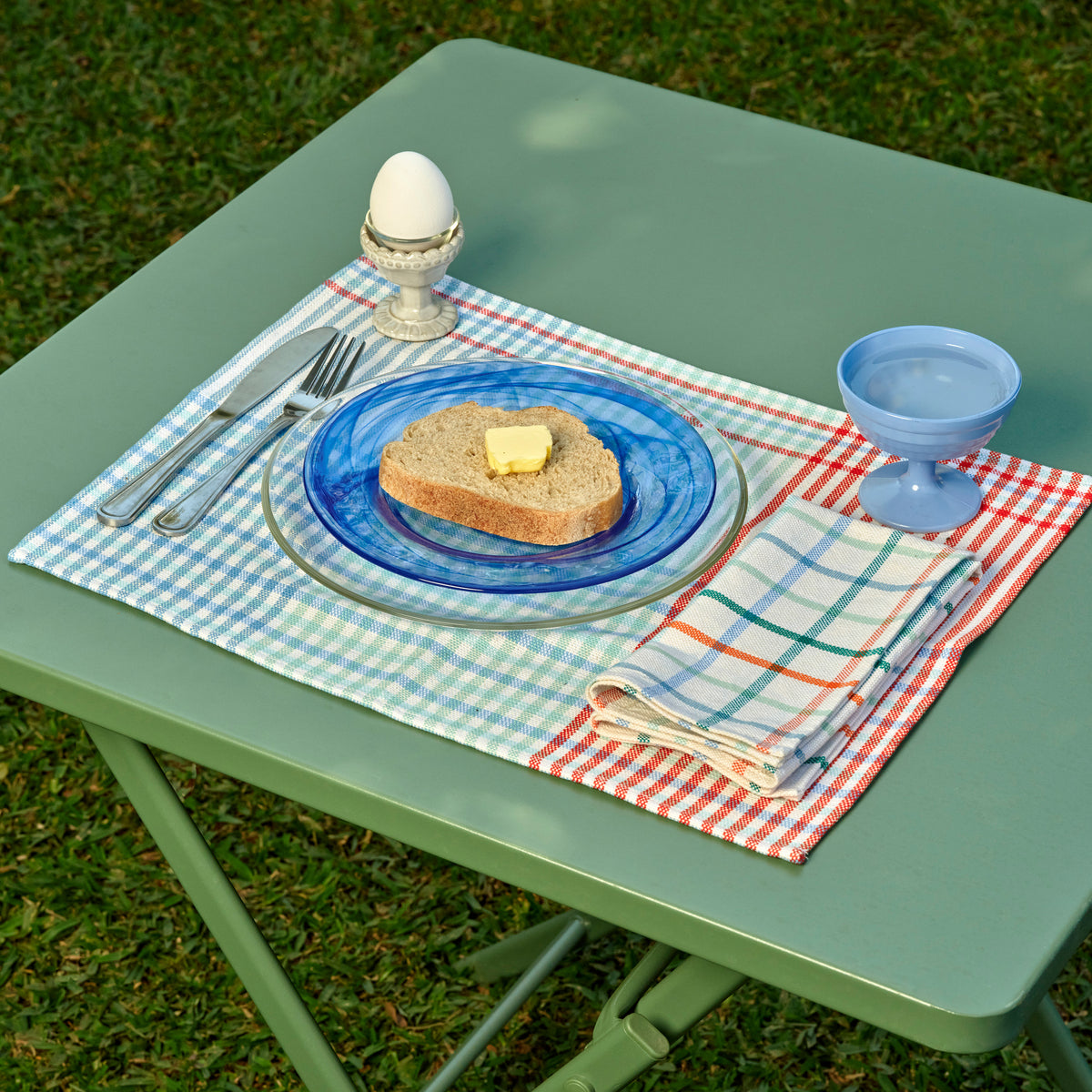 Outdoor picnic-style table setting on a green folding table with a blue-and-white gingham placemat, a blue plate, bread with butter, silver cutlery, a light blue goblet, and a folded striped napkin on grass.