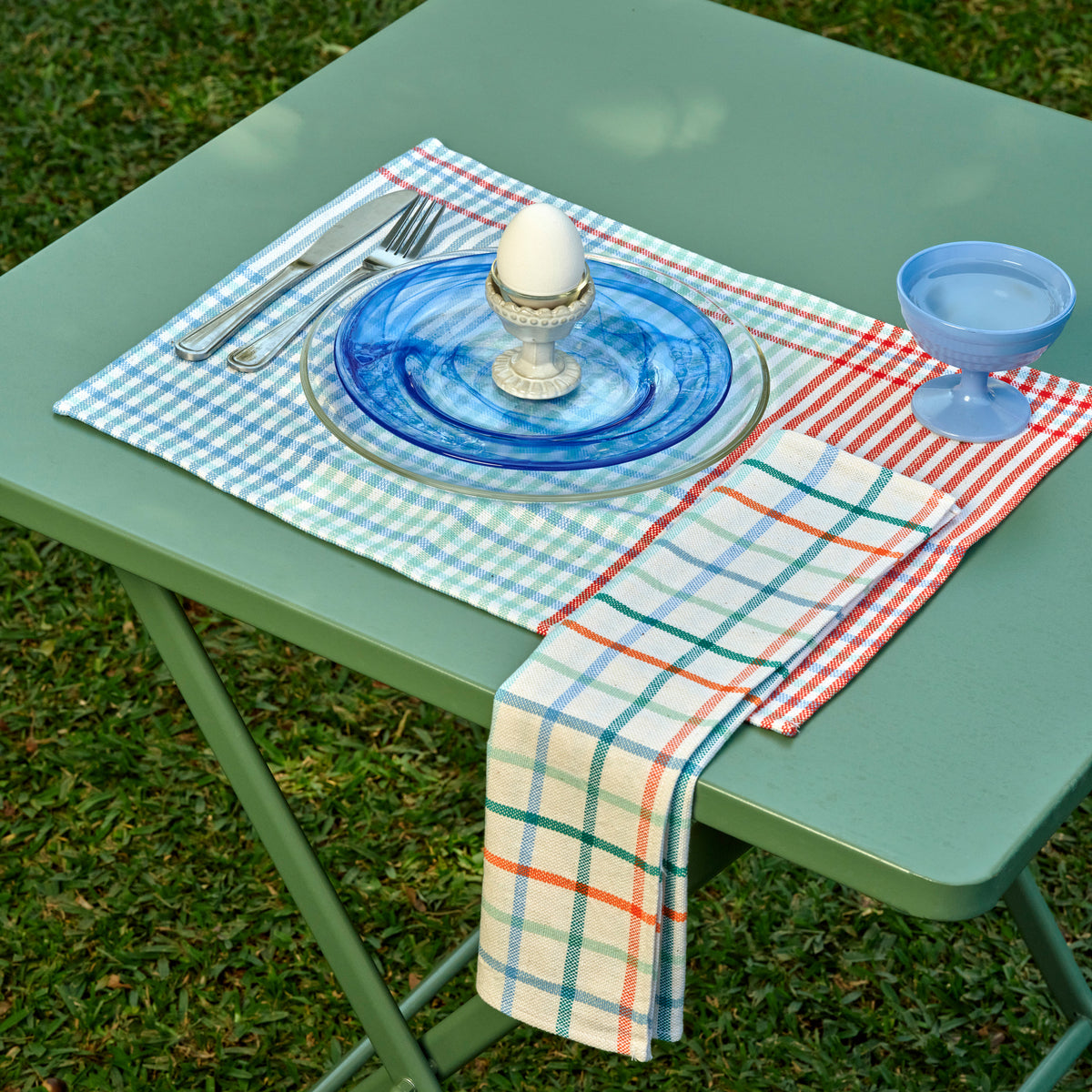 A styled outdoor table setting on a green metal table with grass underneath. The place setting includes a blue glass plate on a clear glass charger, a white egg in a silver egg cup, silver cutlery, a light blue goblet, and folded plaid table linens in blue, red, green, and white.