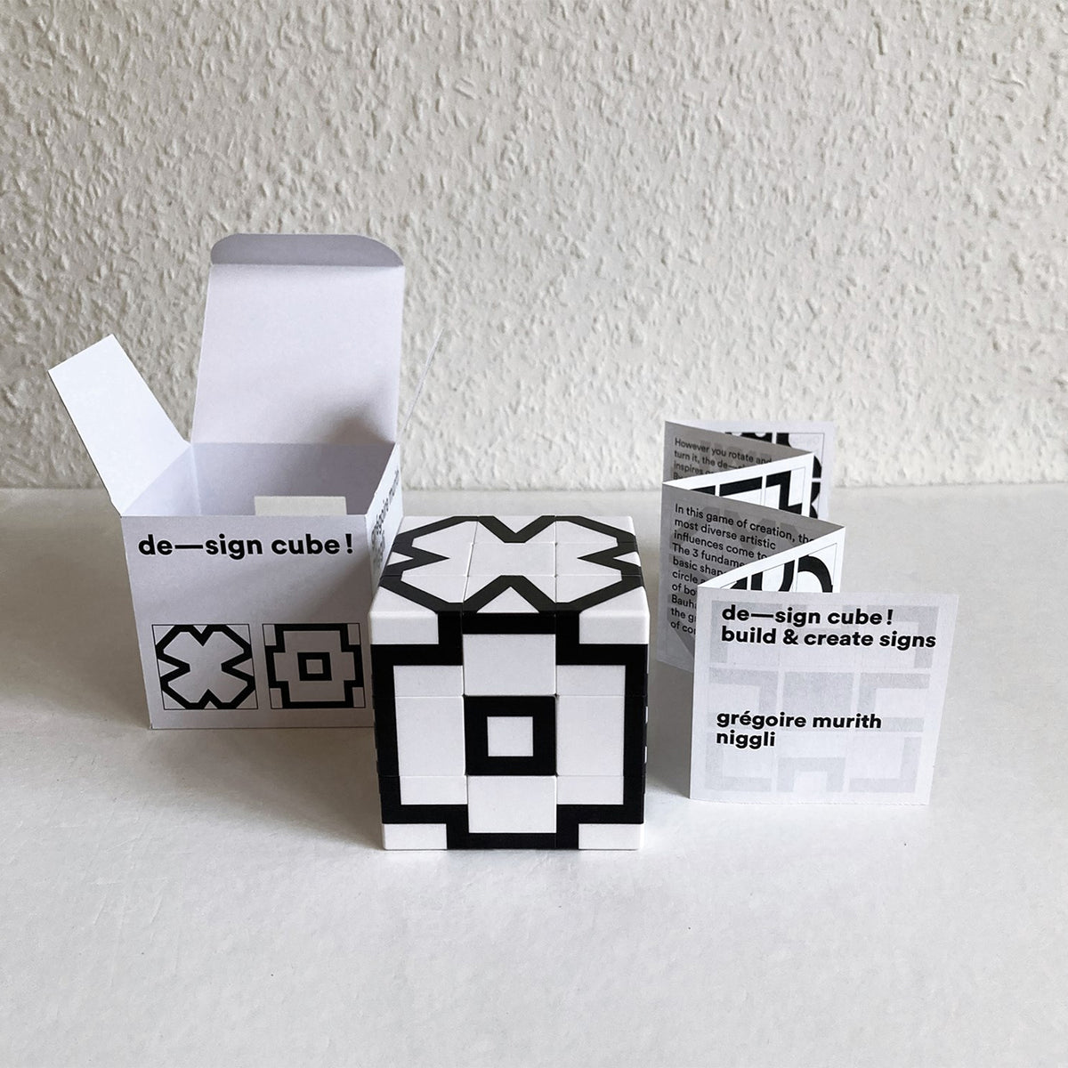 A black and white geometric cube puzzle box with its open white packaging and instruction card on a white surface against a textured white wall.