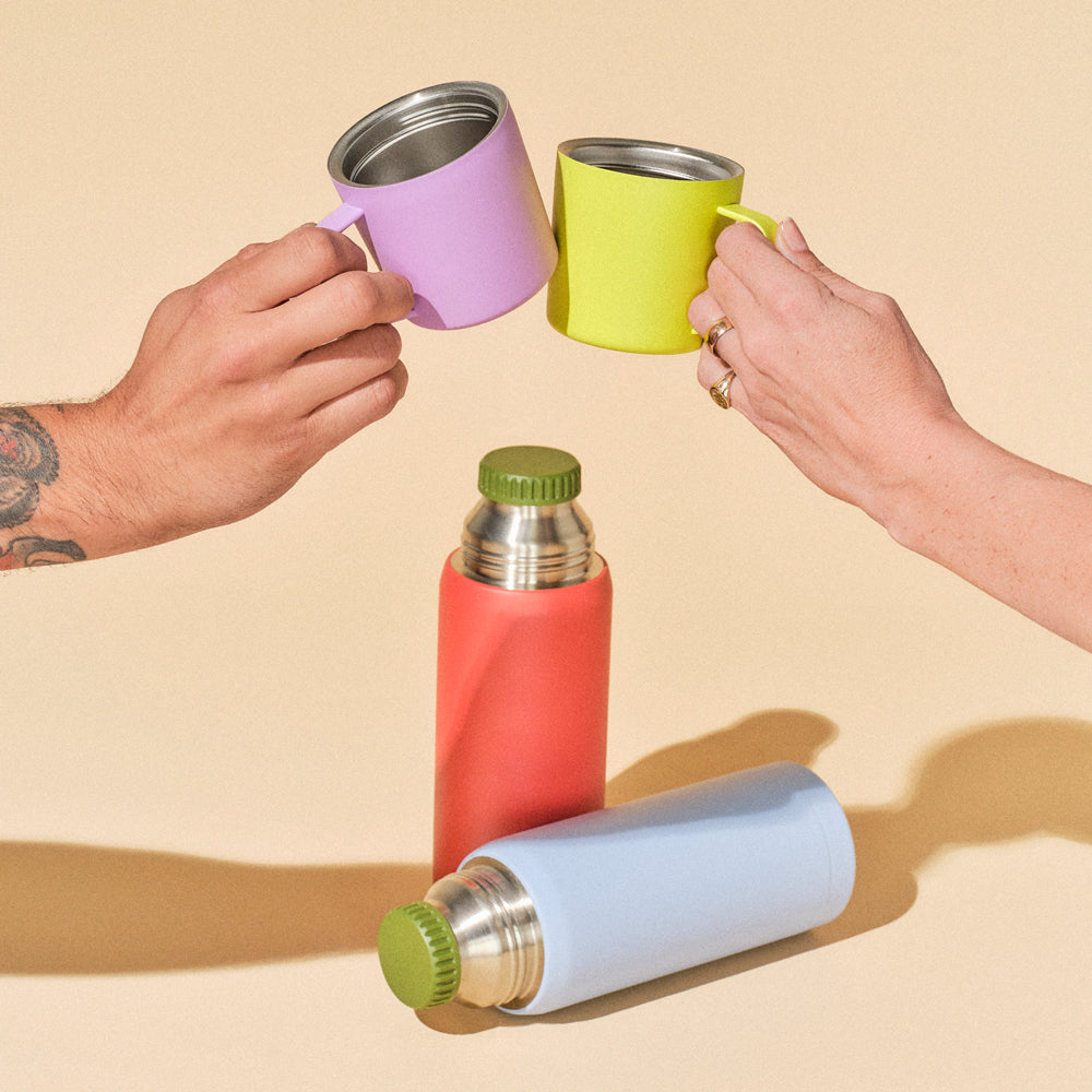 Two hands holding colorful mugs with two bottles on a beige background