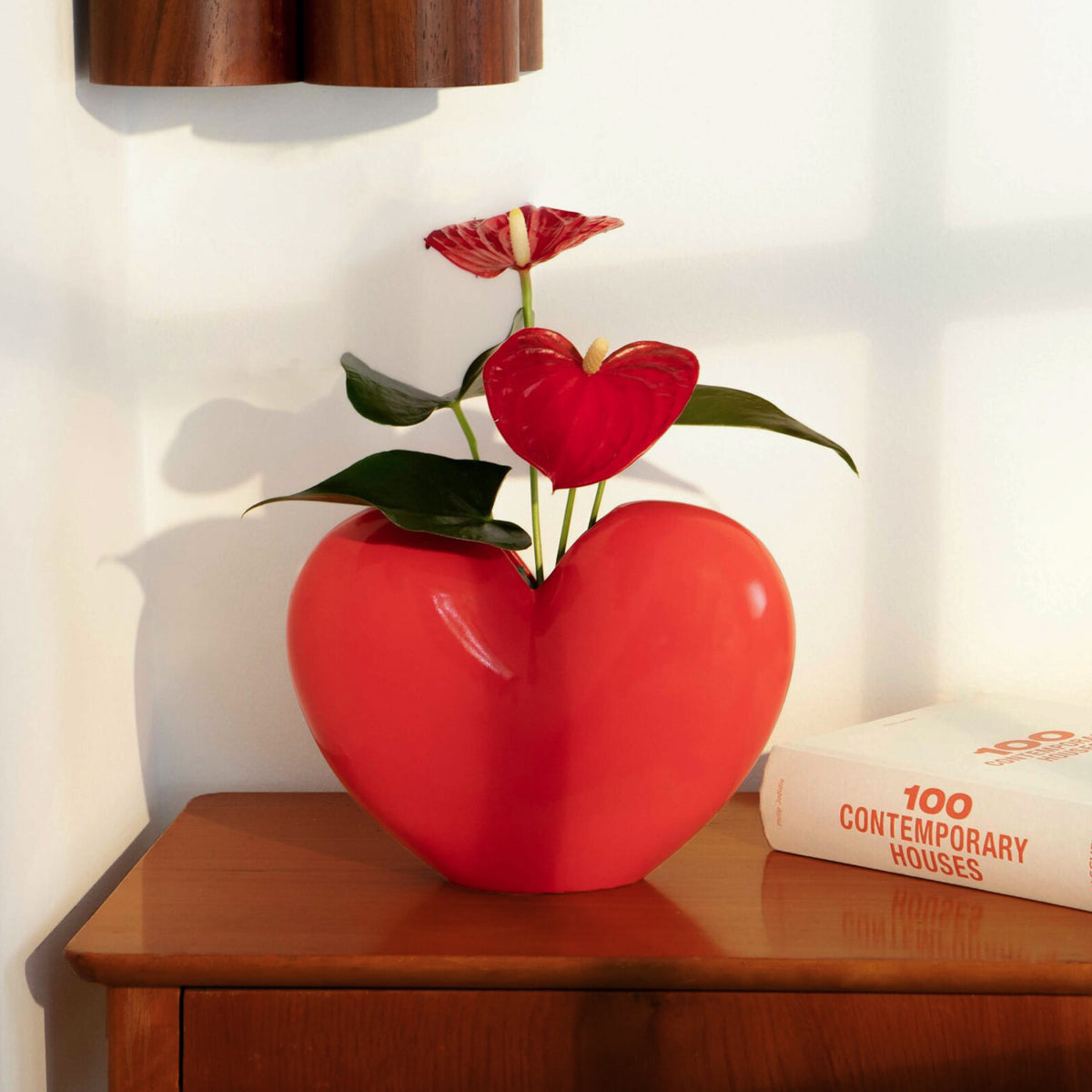 Red heart-shaped vase with a plant on a wooden surface next to a book