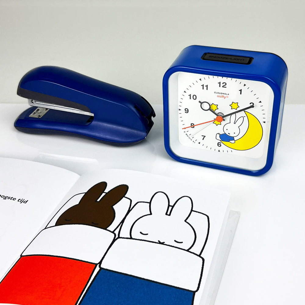 Clock on desk next to stapler and book.