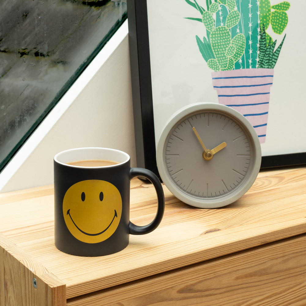 Black mug with yellow smiley face on a wooden surface next to a gray clock and framed picture of cacti.