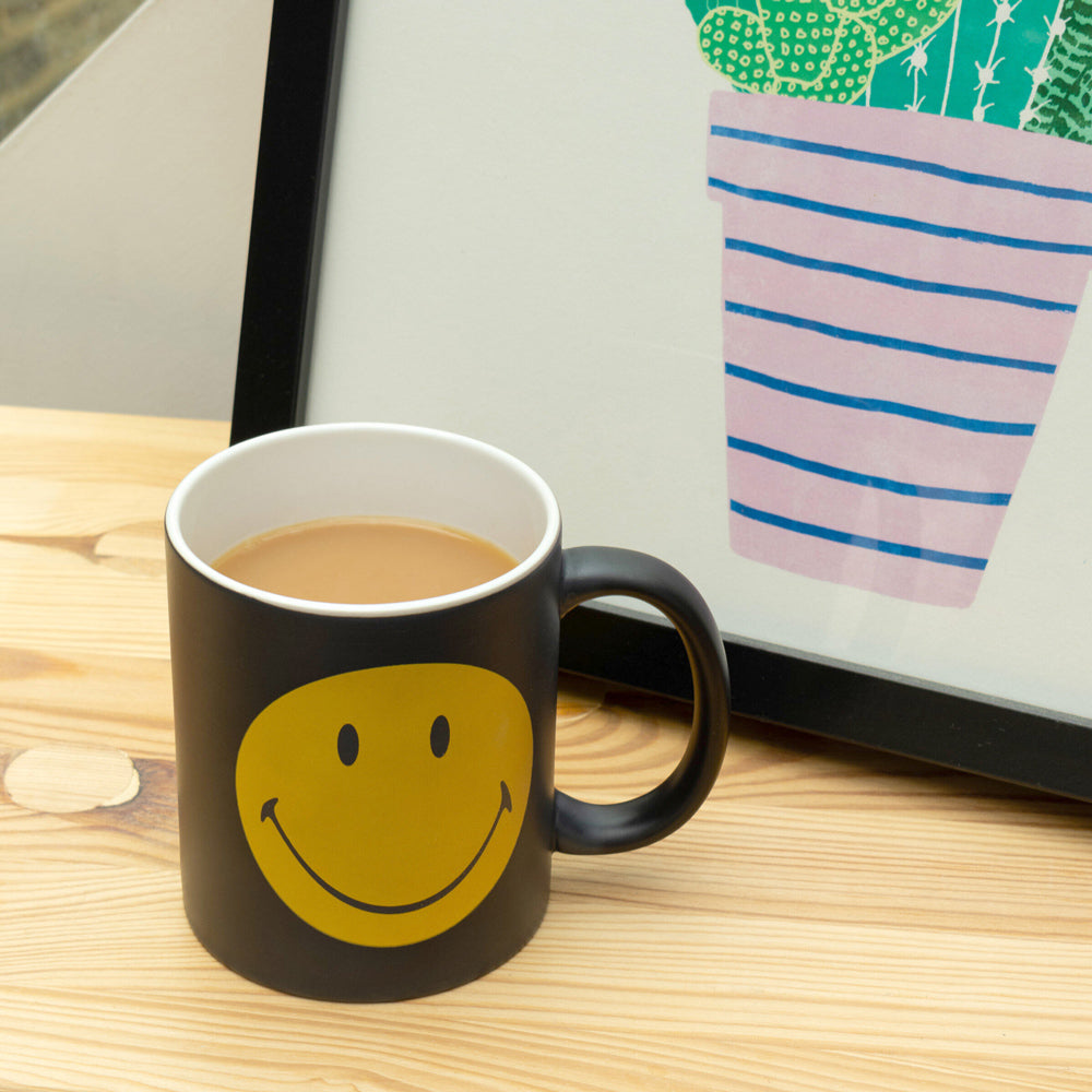 Black mug with yellow smiley face on a wooden surface next to a laptop.