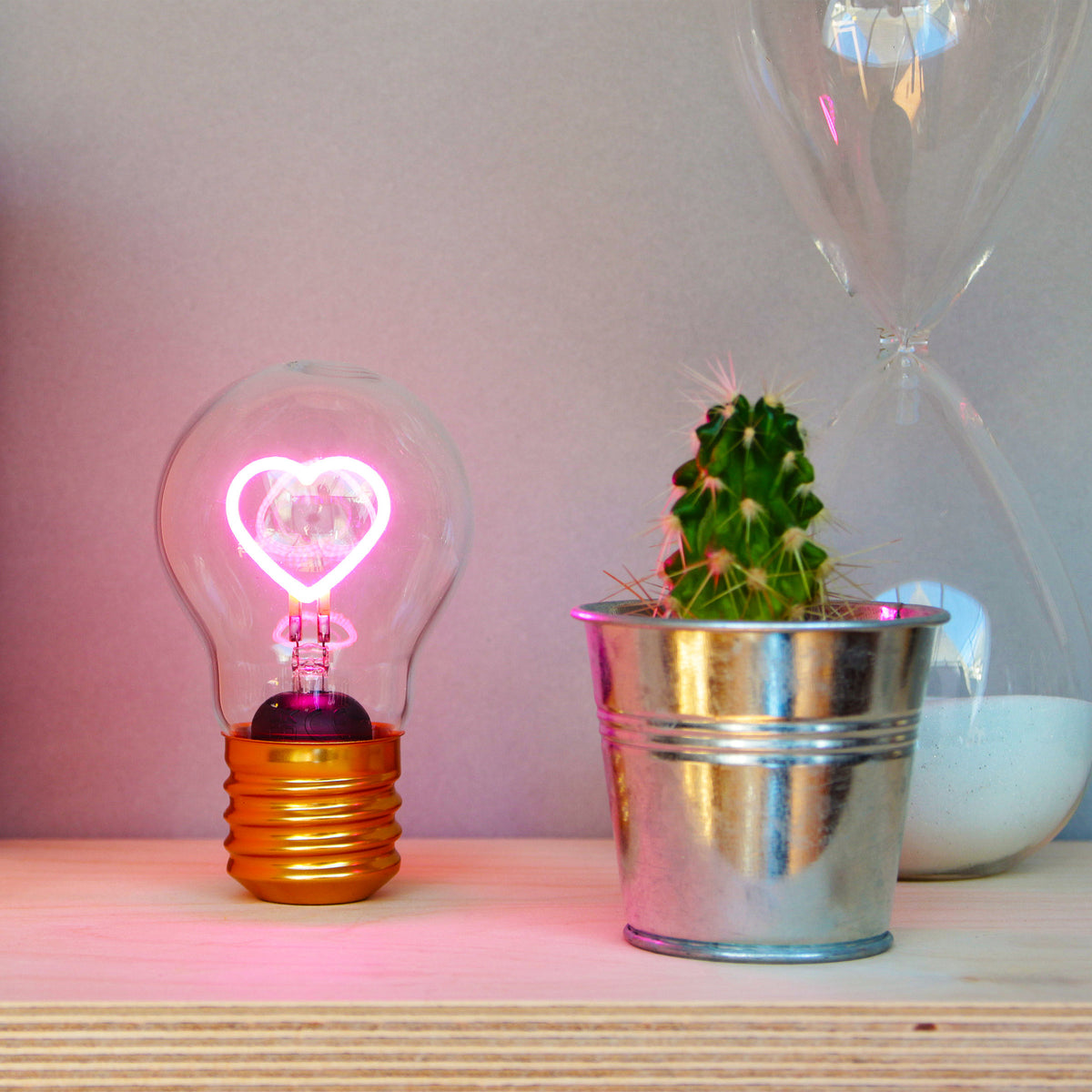 Lightbulb with heart-shaped LED filament, on shelf with cactus and hourglass.