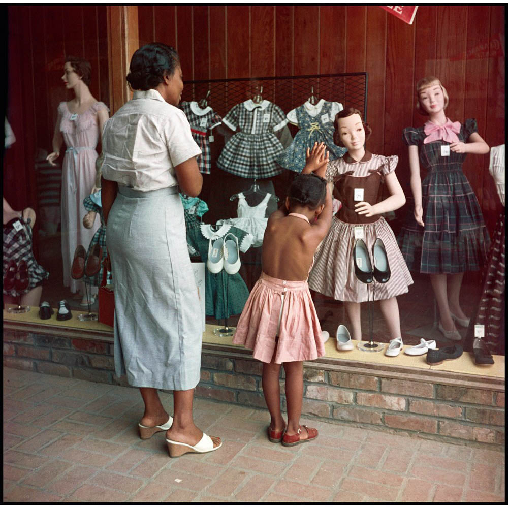 Black mother and daughter look at white mannequins wearing dresses in shop window.