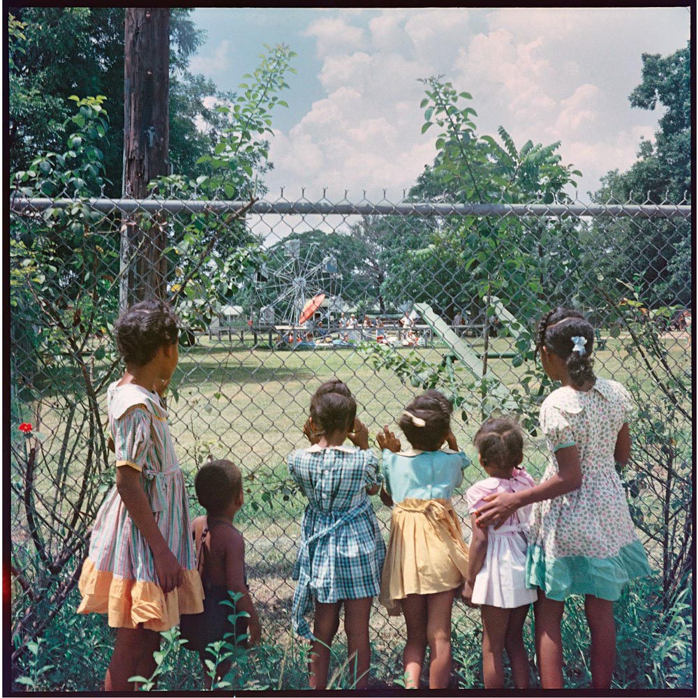 Black children looking at carnival rides through a chain link fence.