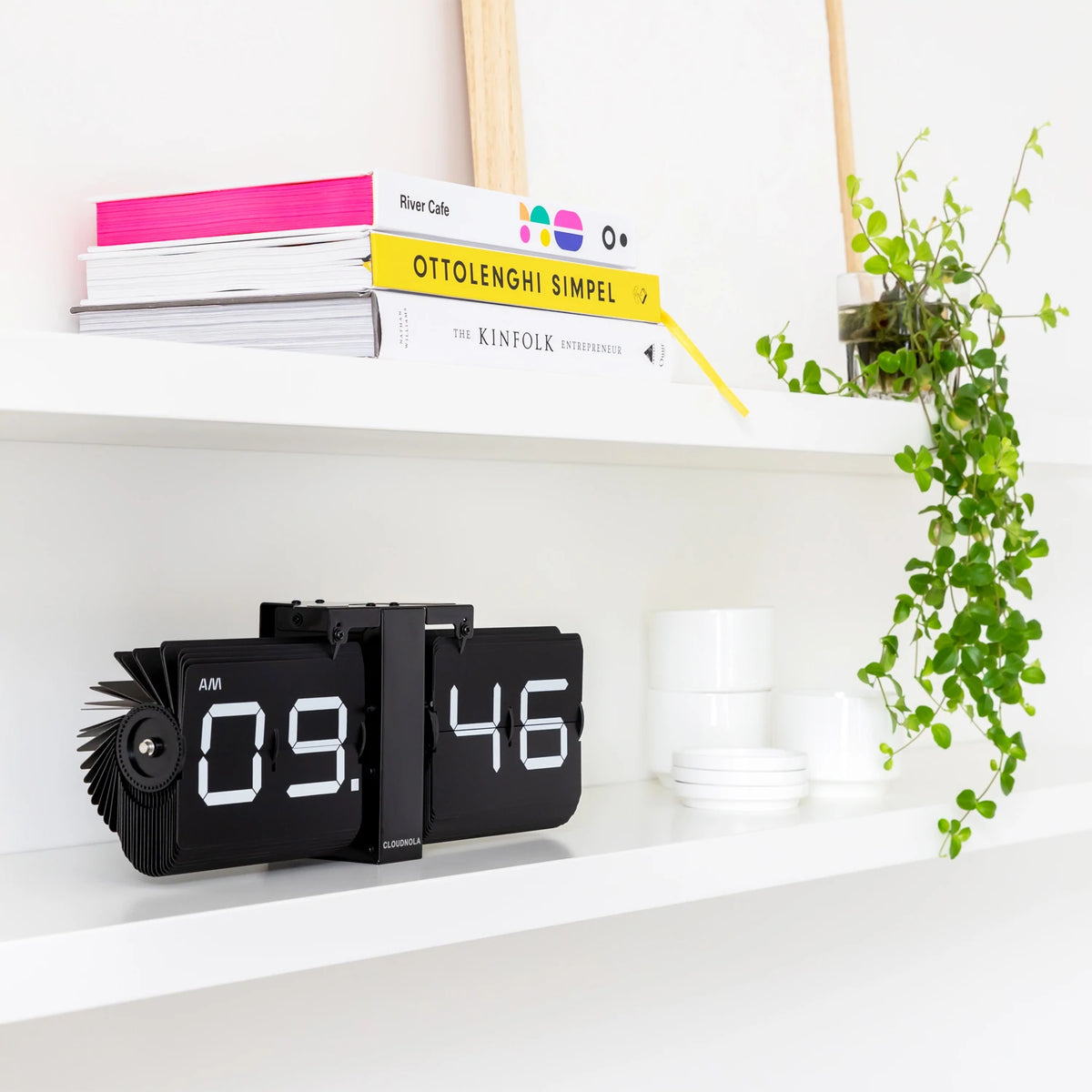 The Flipping Out Clock: Black displayed on a shelf with books and a plant.