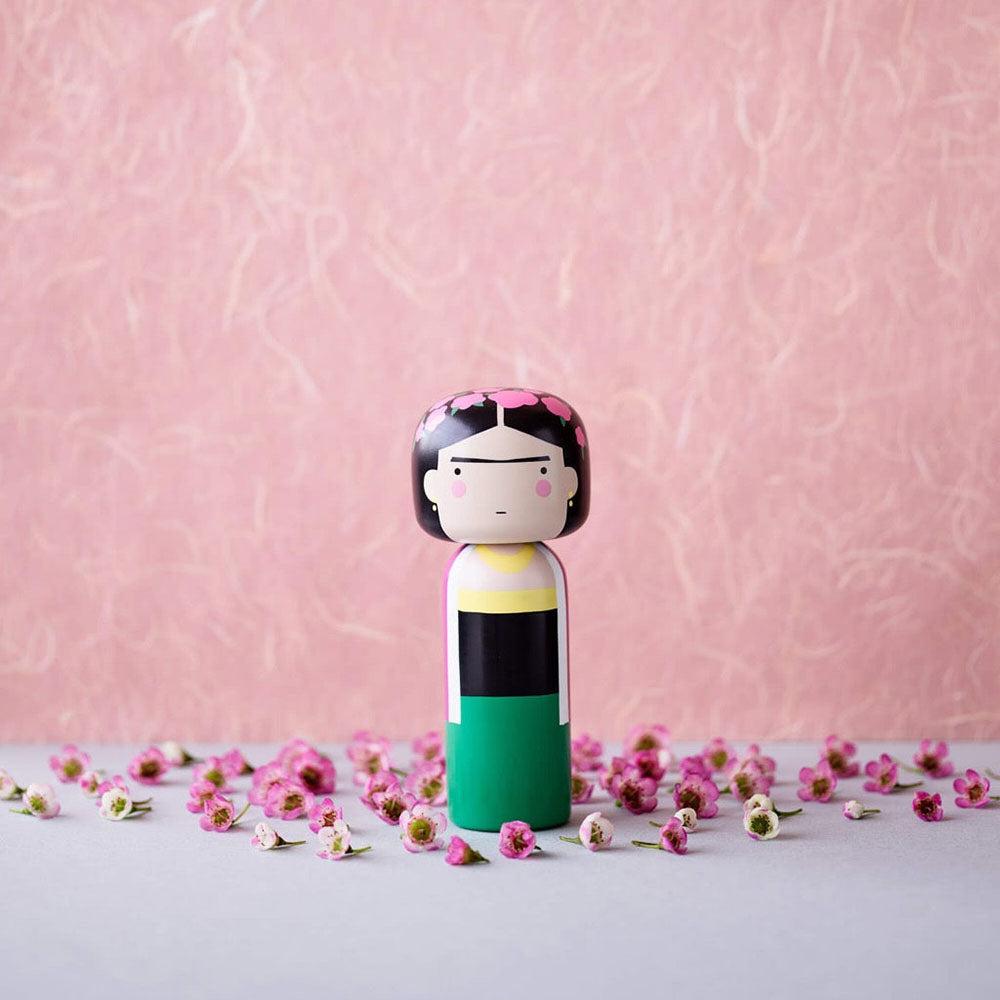 The Frida Kokeshi Doll on display with miniature flowers and a textured pink background.