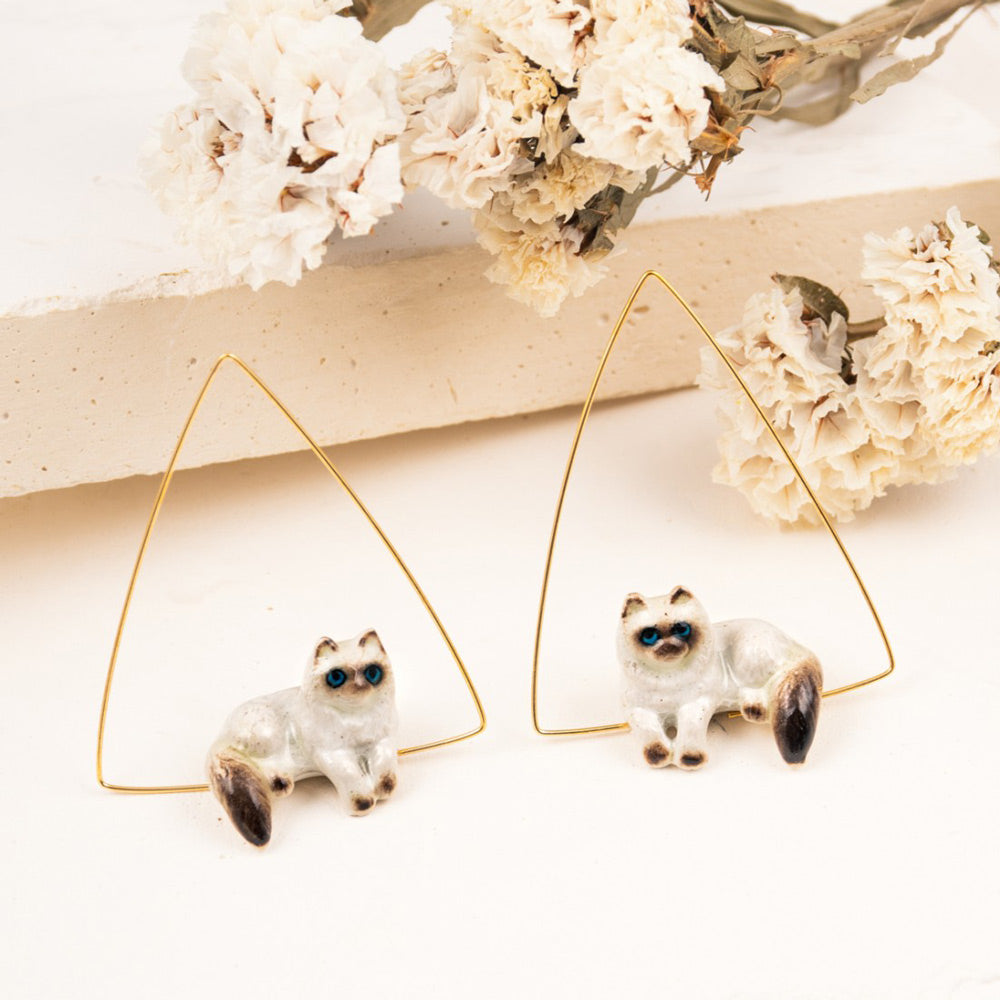 Siamese Cat Triangle Earrings by Nach on white plaster and floral decor.